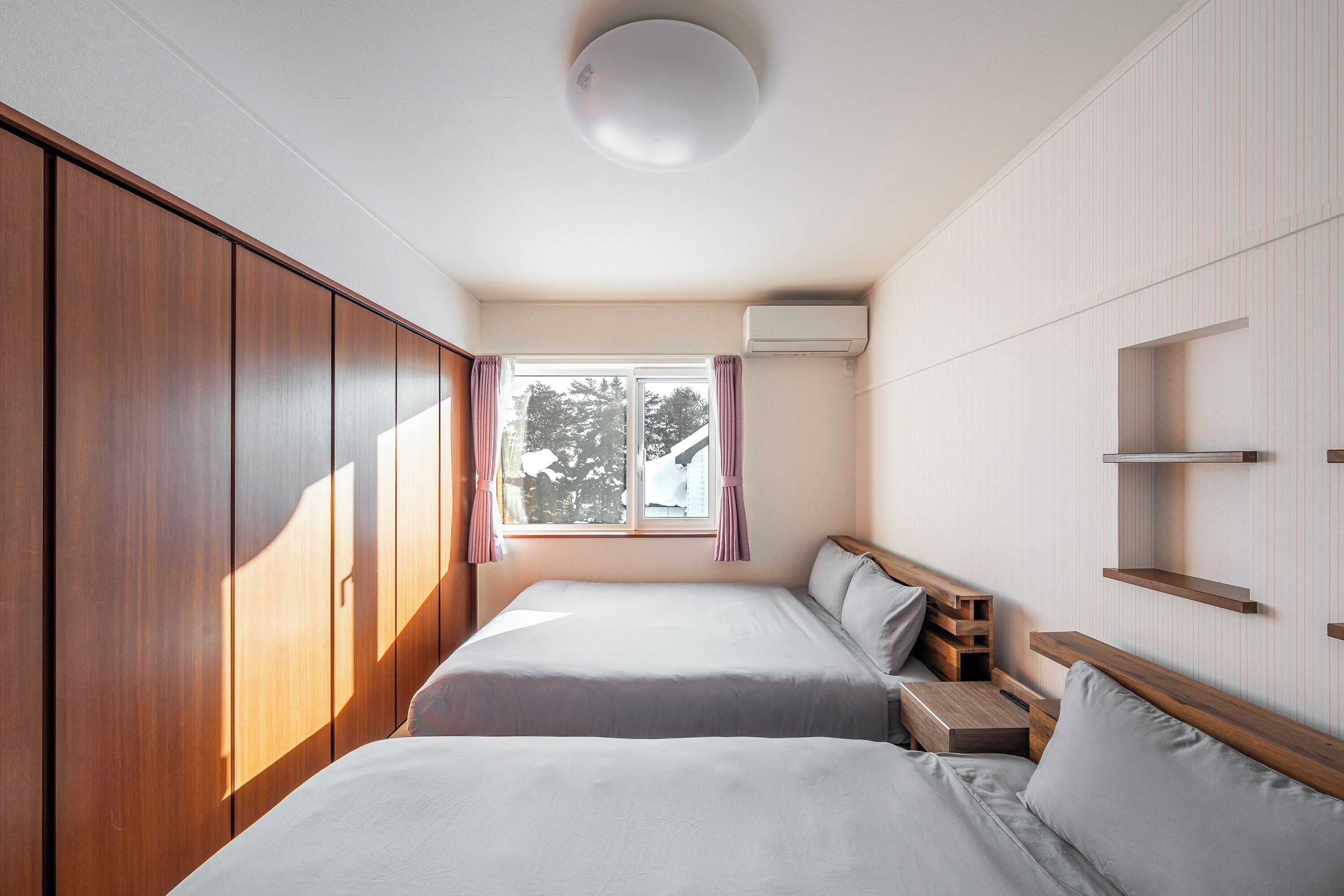 Une chambre avec deux lits, une armoire en bois, une fenêtre avec des rideaux et des étagères intégrées.