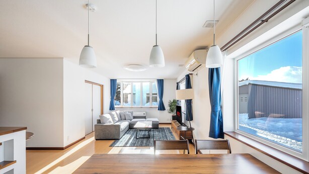 Un salon moderne avec une table à manger en bois, un canapé et un téléviseur. De grandes fenêtres, dotées de rideaux, offrent une vue sur l’extérieur.