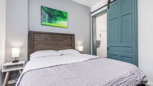 A neatly made bed with a wooden headboard, a bedside table with a lamp, and a door with a towel hanging on it. - 1103#203 / Downtown Oasis: En-Suite Master (Dubuque)