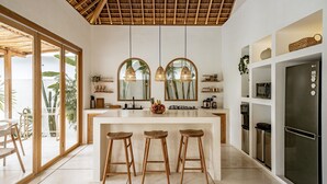 A modern kitchen with a central island, wooden stools, a refrigerator, and a view of the ocean through large windows. - Villa KerangSue (Canggu)