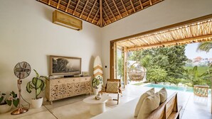 A modern living room with a TV, sofa, and a view of a swimming pool. - Villa KerangSue (Canggu)