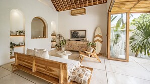 A modern living room with a wooden bench, a sofa, a TV, and a glass door leading to a garden. - Villa KerangSue (Canggu)