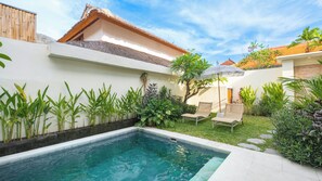 A swimming pool with a white wall and tropical plants, a wooden fence, and a thatched-roof building. - Villa Ambalika by Villa Finder (Seminyak)
