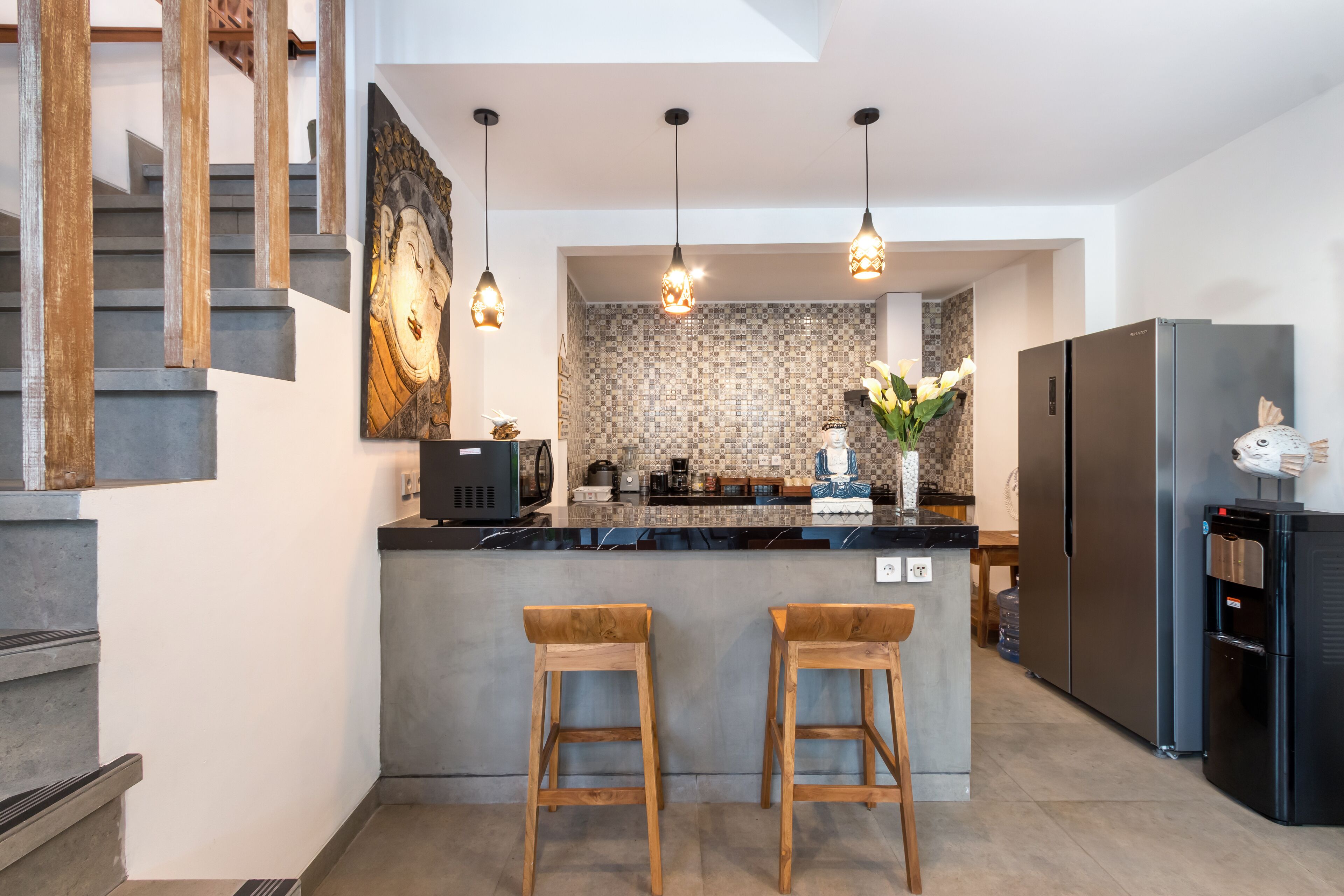 A modern kitchen with a central island, wooden stools, hanging pendant lights, and a stainless steel refrigerator.