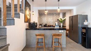 Uma cozinha moderna com uma ilha central, banquetas de madeira, luminárias pendentes e uma geladeira de aço inoxidável.