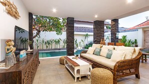 Ampla sala de estar com piscina, móveis de madeira e plantas tropicais.
