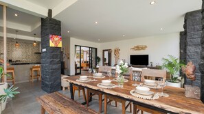 Un coin repas avec une table en bois dressée pour quatre personnes, entouré de piliers en pierre, une cuisine en arrière-plan et un salon avec un téléviseur et des plantes.