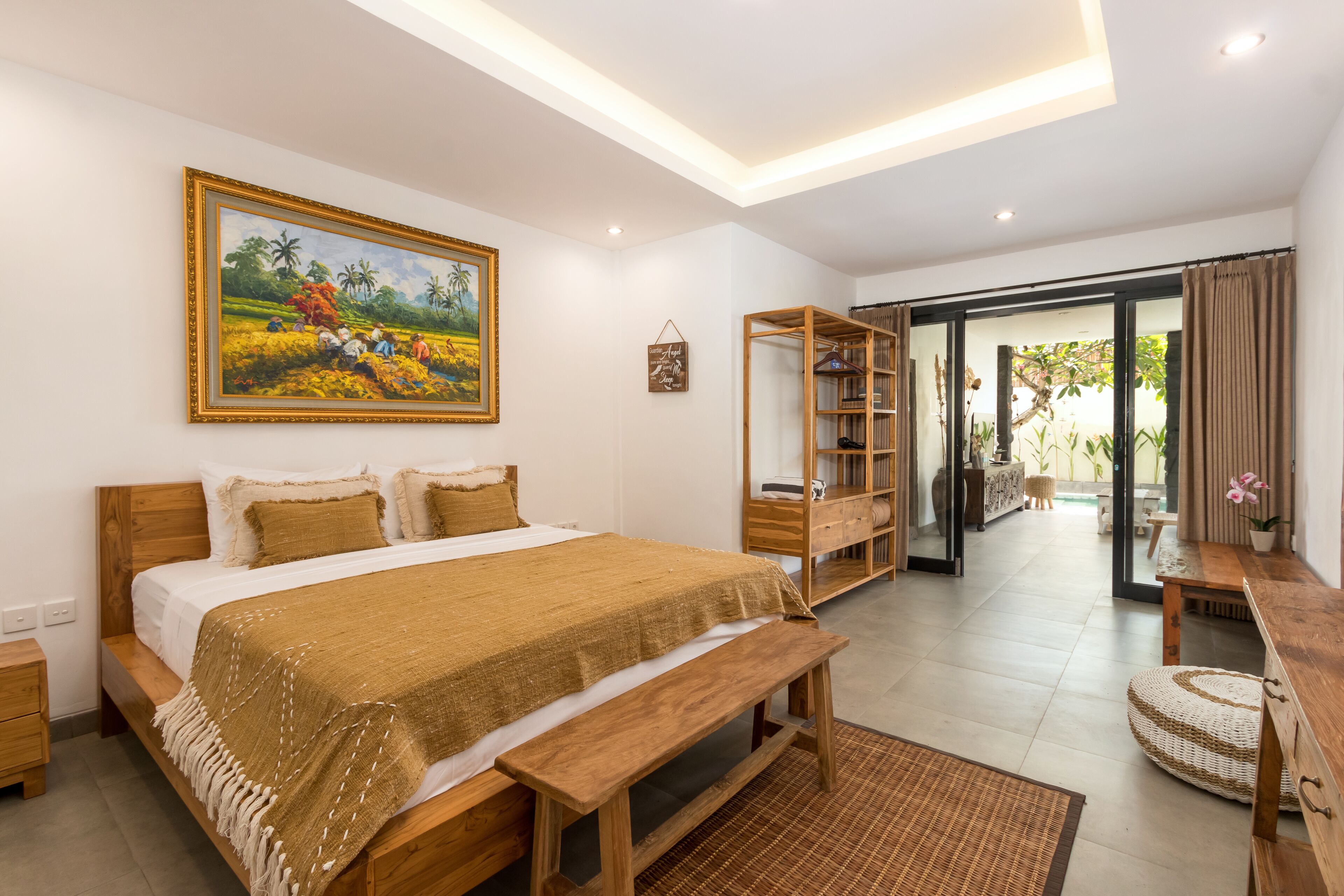 A bedroom with a large bed, wooden headboard, a bench, a bookshelf, and a painting on the wall.