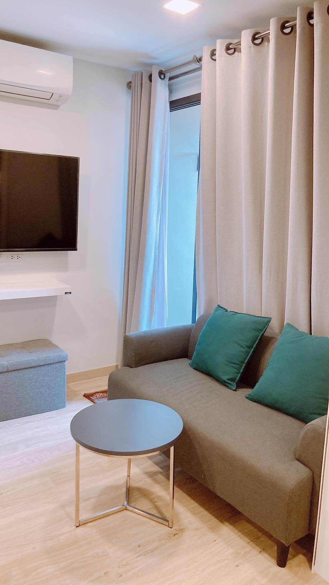 A modern living room with a grey sofa, green cushions, a small round table, and a flat-screen TV mounted on the wall.