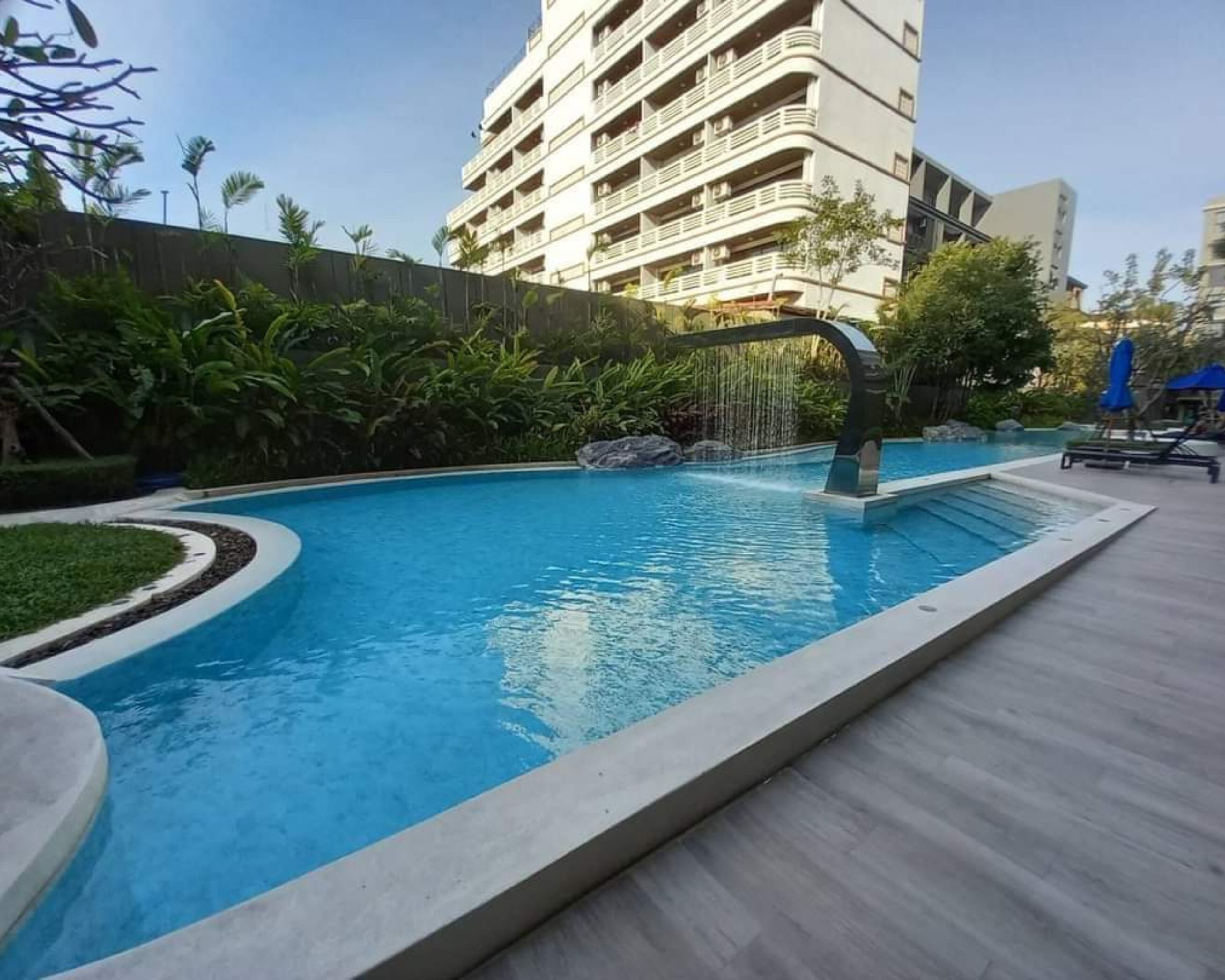 Une piscine agrémentée d’une cascade, entourée d’un jardin aménagé et avec un immeuble à plusieurs étages en arrière-plan.