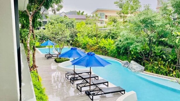 A swimming pool with lounge chairs and umbrellas, surrounded by greenery and a building in the background.