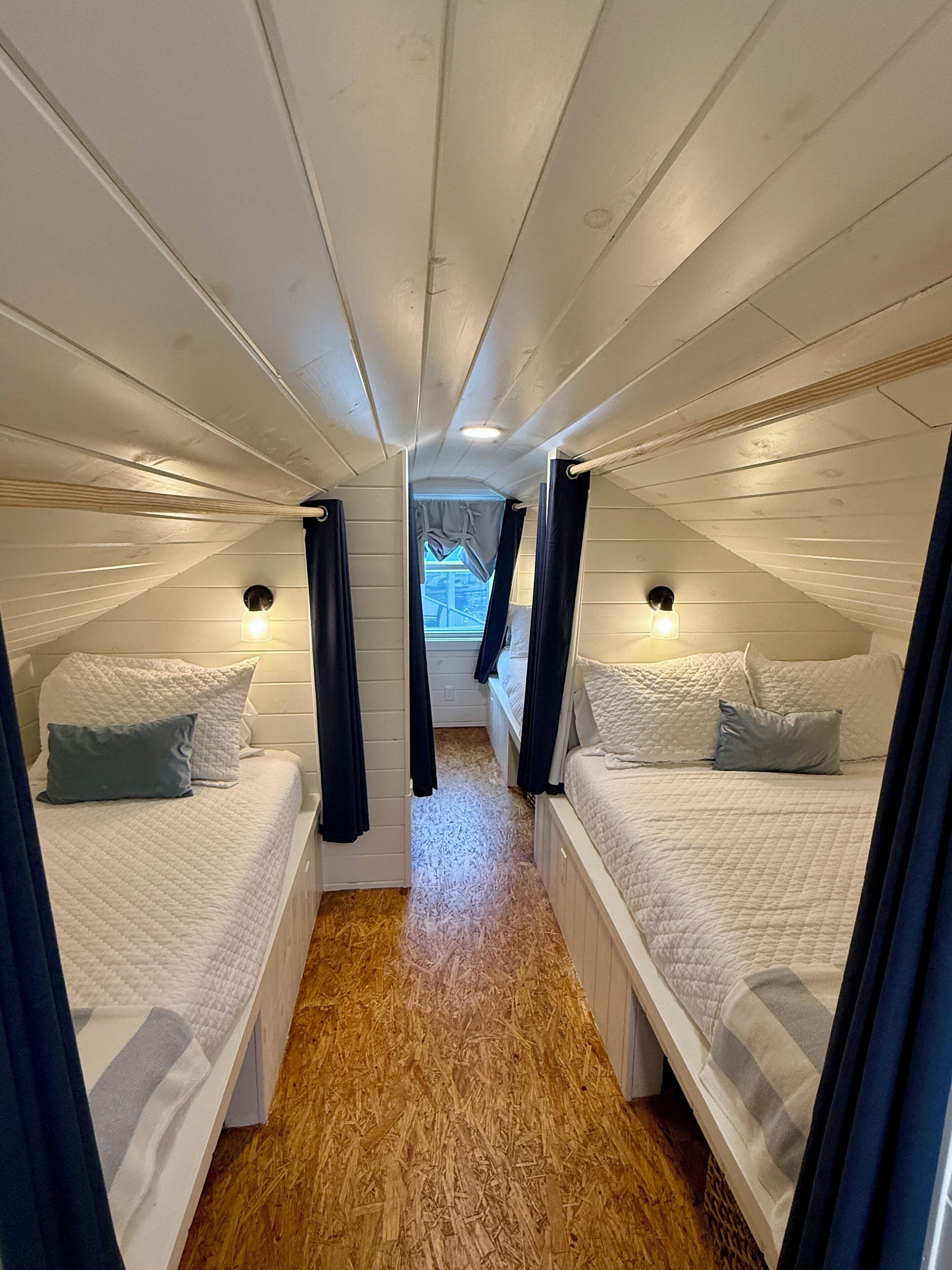 Una habitación pequeña y cerrada con dos camas, cada una con almohada y cabecera. El techo es inclinado, revestido de paneles de madera, y al fondo hay una ventana pequeña.