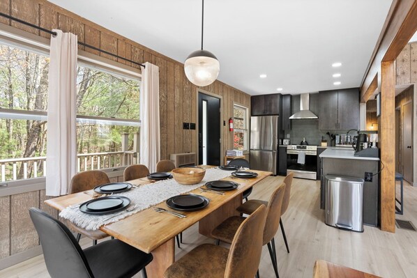 Una cocina moderna con una isla central, un comedor con una mesa larga preparada para cuatro personas, y ventanas amplias con cortinas.