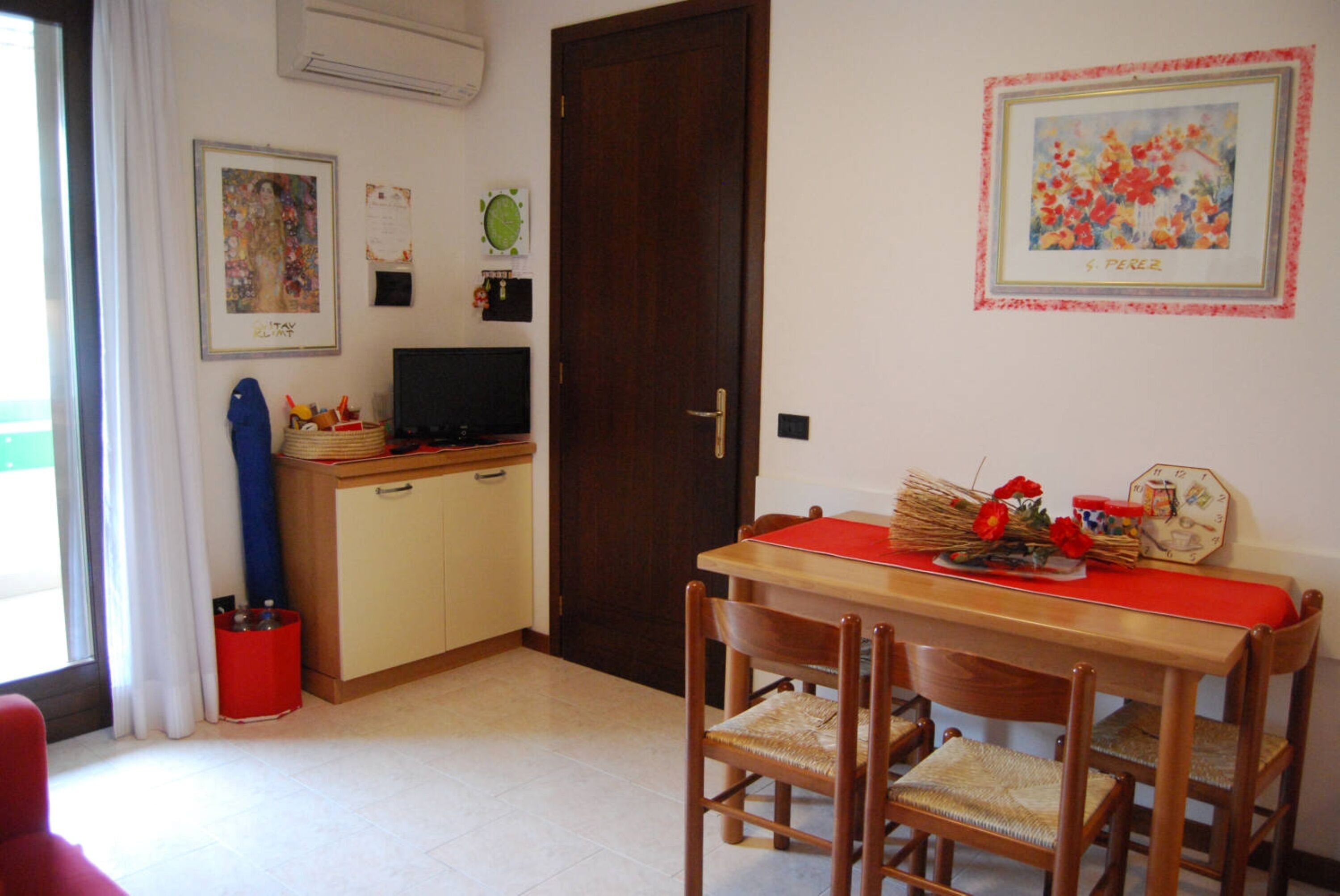 A dining area with a wooden table, four chairs, and a red table runner. There is a television on a cabinet, a clock on the wall, and a framed picture above the table.