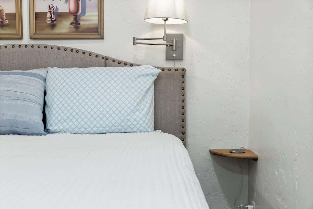 Un dormitorio con una cama, dos almohadas, una lámpara de pared y una mesita de noche de madera.