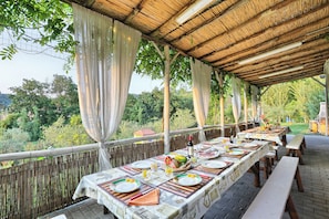 An outdoor dining area with tables set for a meal, surrounded by a bamboo fence, and overlooking a lush garden.