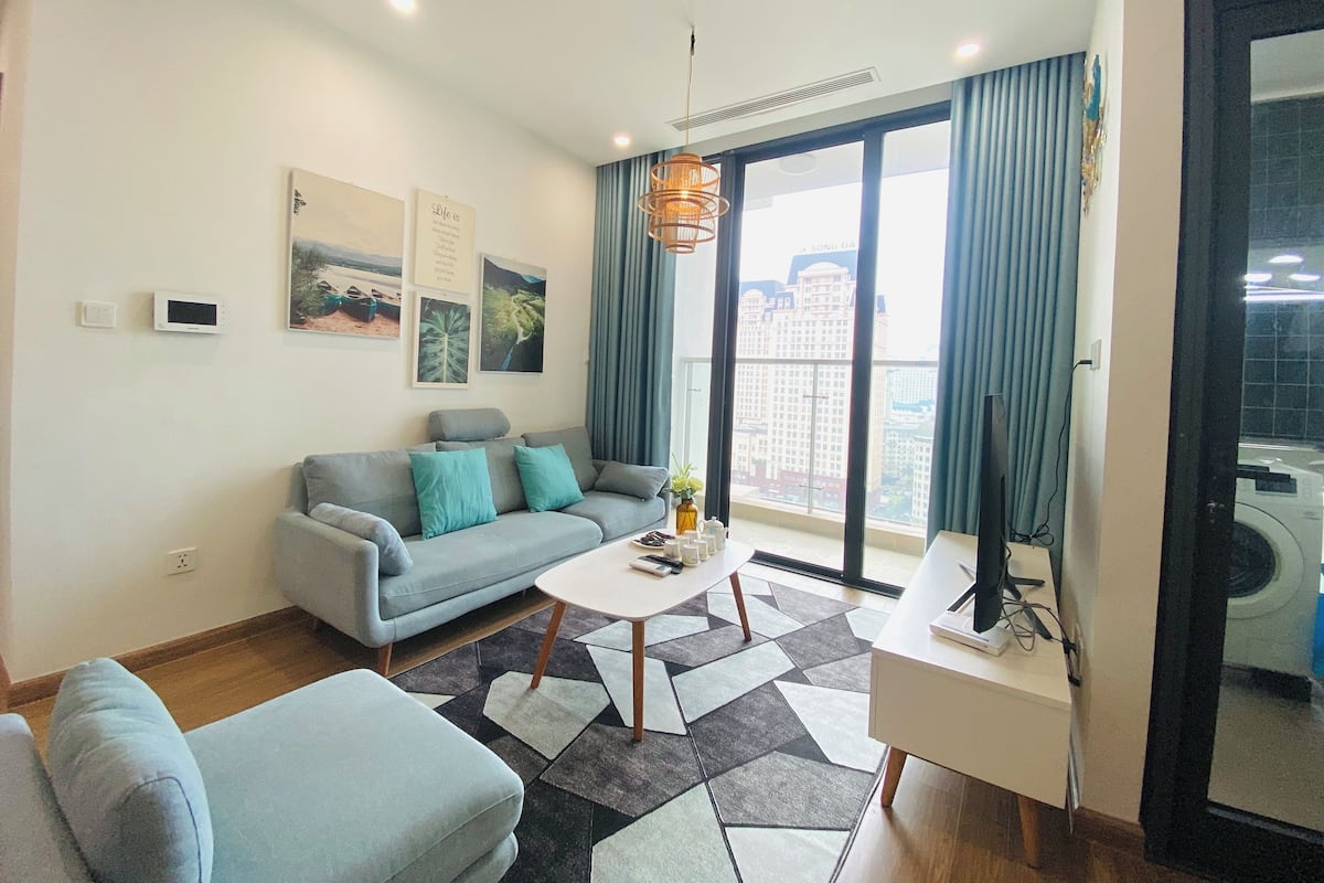 Ein modernes Wohnzimmer mit grauem Sofa, einem Couchtisch und Blick auf eine Stadtkulisse durch große Fenster.