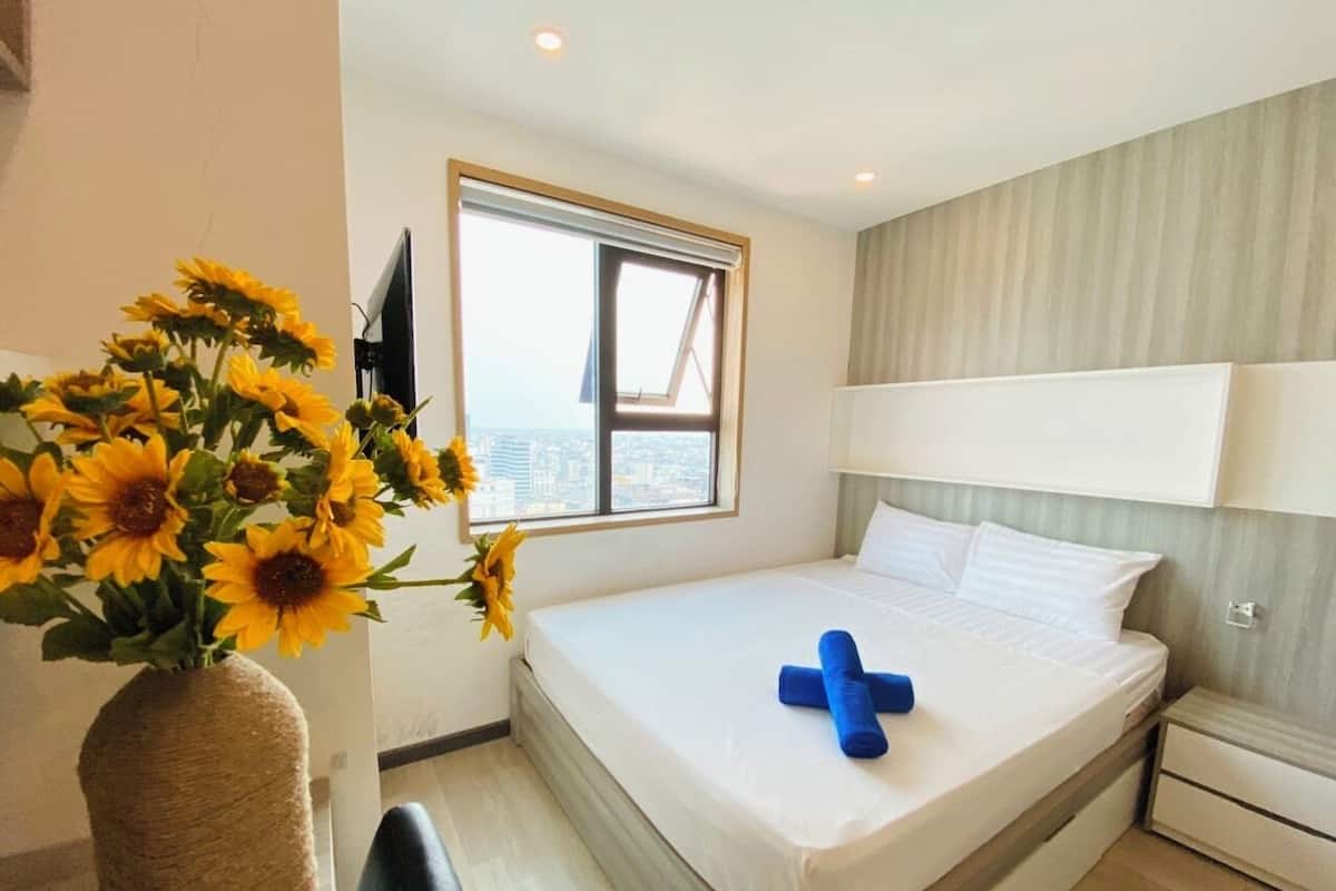 A hotel room with a bed, a nightstand, a lamp, a window with a view, and a vase of sunflowers.