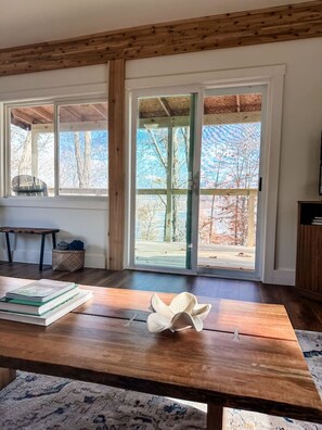 Een kamer met een houten tafel, boeken, een decoratief object, een bank en uitzicht door grote ramen.