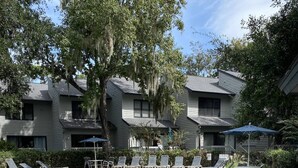 A pool area with lounge chairs and umbrellas, surrounded by trees and a building with multiple windows. - Treetops 4502 - an iTrip Hilton Head Home (Hilton Head Island)