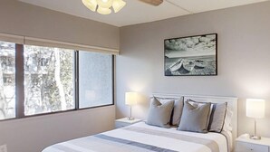 A bedroom with a bed, two bedside tables, a ceiling fan, a window, and a wall-mounted picture. - Treetops 4502 - an iTrip Hilton Head Home (Hilton Head Island)