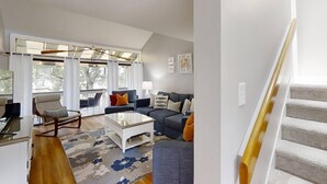 A spacious living room with a large window, a sofa set, a coffee table, and a staircase. - Treetops 4502 - an iTrip Hilton Head Home (Hilton Head Island)