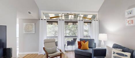 A modern living room with a sofa, armchair, and a TV. There is a large window with curtains, a ceiling fan, and a lamp.