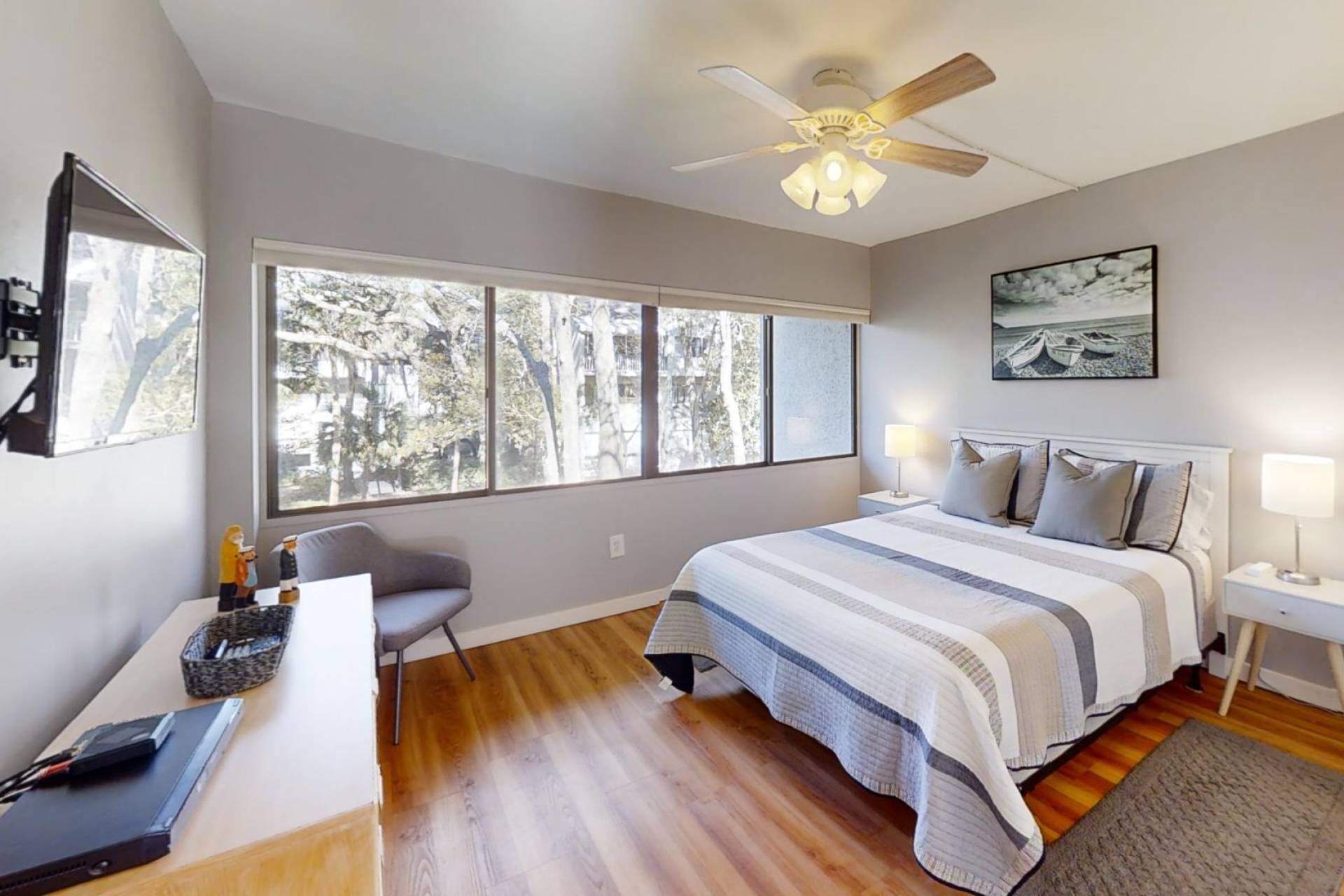 A bedroom with a large window, a bed with a striped blanket, a desk with a laptop, and a chair.
