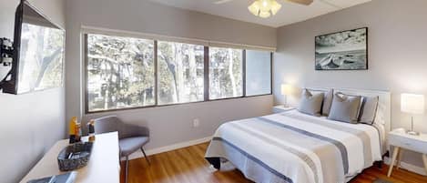 A bedroom with a large window, a bed with a striped blanket, a desk with a laptop, and a chair.