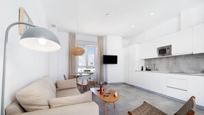 A modern living room with a sofa, a wooden chair, a dining table with a wine bottle, and a kitchen area with white cabinets and a microwave.
