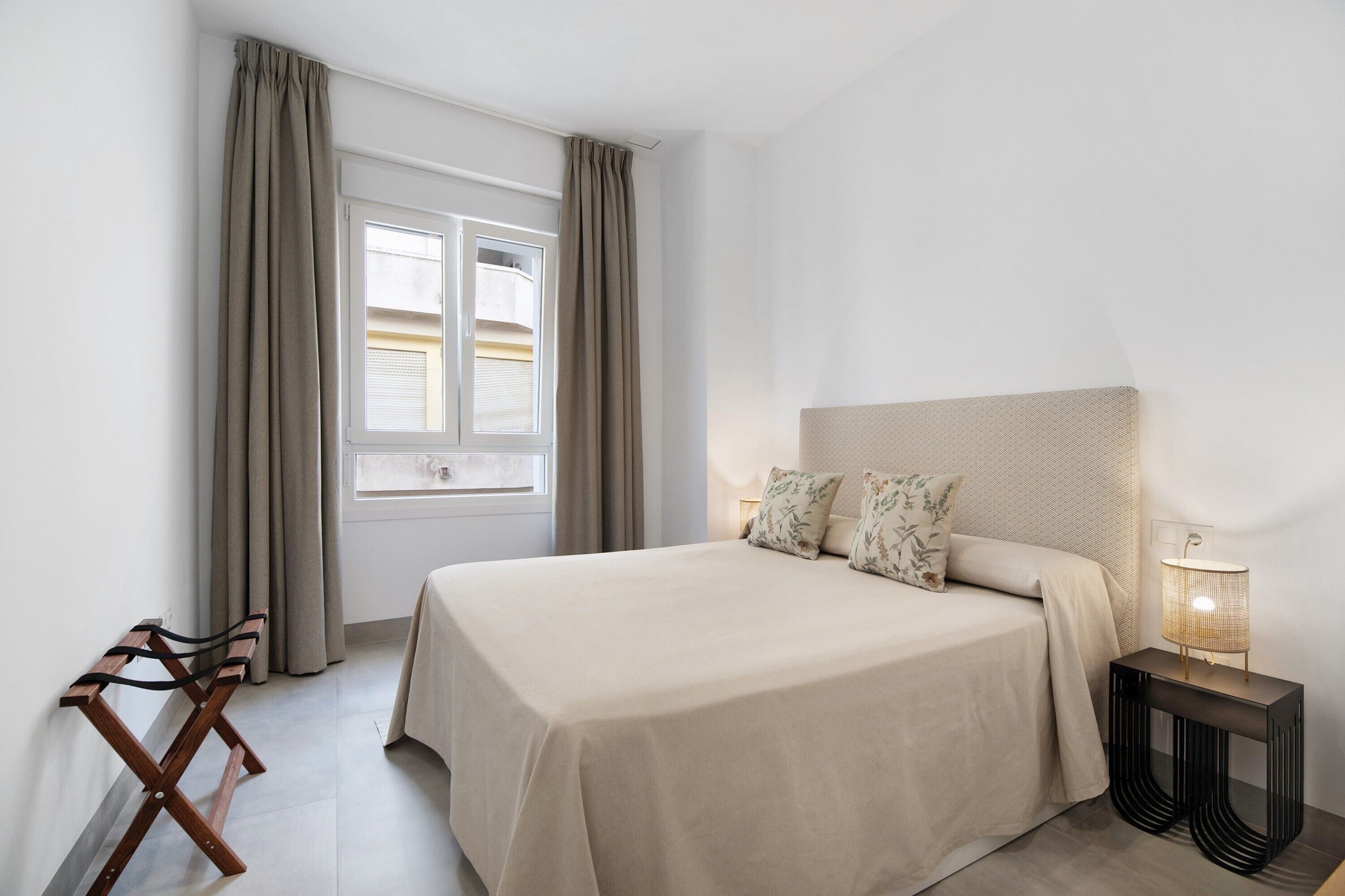 Een slaapkamer met een bed, een nachtkastje met een lamp, een raam met gordijnen en een houten bankje.