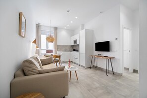 A modern living room with a sofa, a dining area with chairs, a kitchen with a microwave, and a TV on the wall.