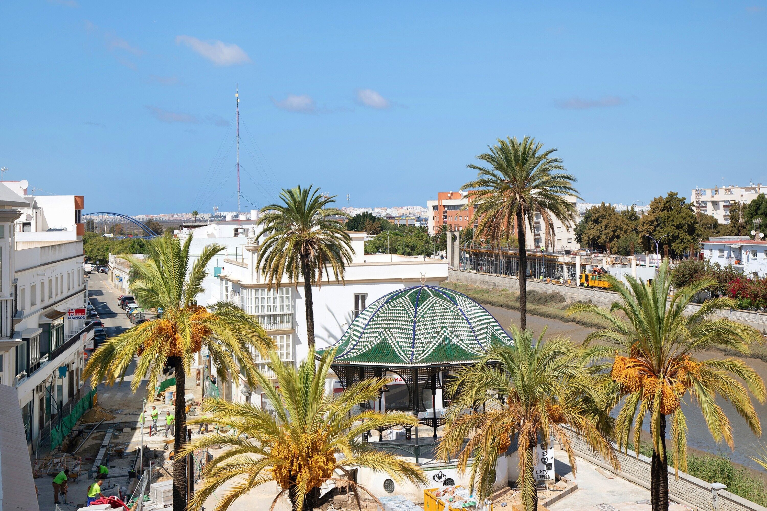 Une vue sur un paysage urbain avec des palmiers, un kiosque à toit végétalisé et des bâtiments en arrière-plan.