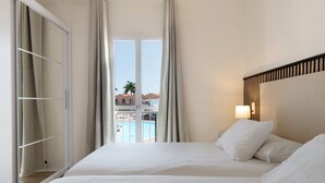Une chambre d’hôtel avec un lit, une fenêtre avec des rideaux, une vue sur une piscine et des bâtiments, et une table de chevet avec une lampe.
