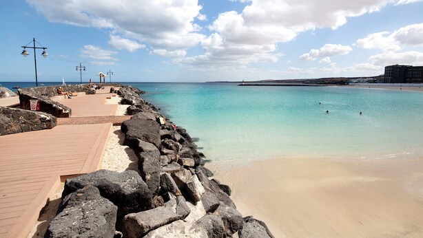 Un paseo marítimo con una barrera rocosa, una playa arenosa y un cielo azul despejado.