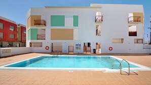 Una piscina con agua cristalina azul, rodeada por una terraza con baldosas y un edificio blanco con balcones y ventanas al fondo.