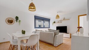 Una sala de estar moderna con sofá, mesa de comedor, sillas, televisor y lámparas colgantes.