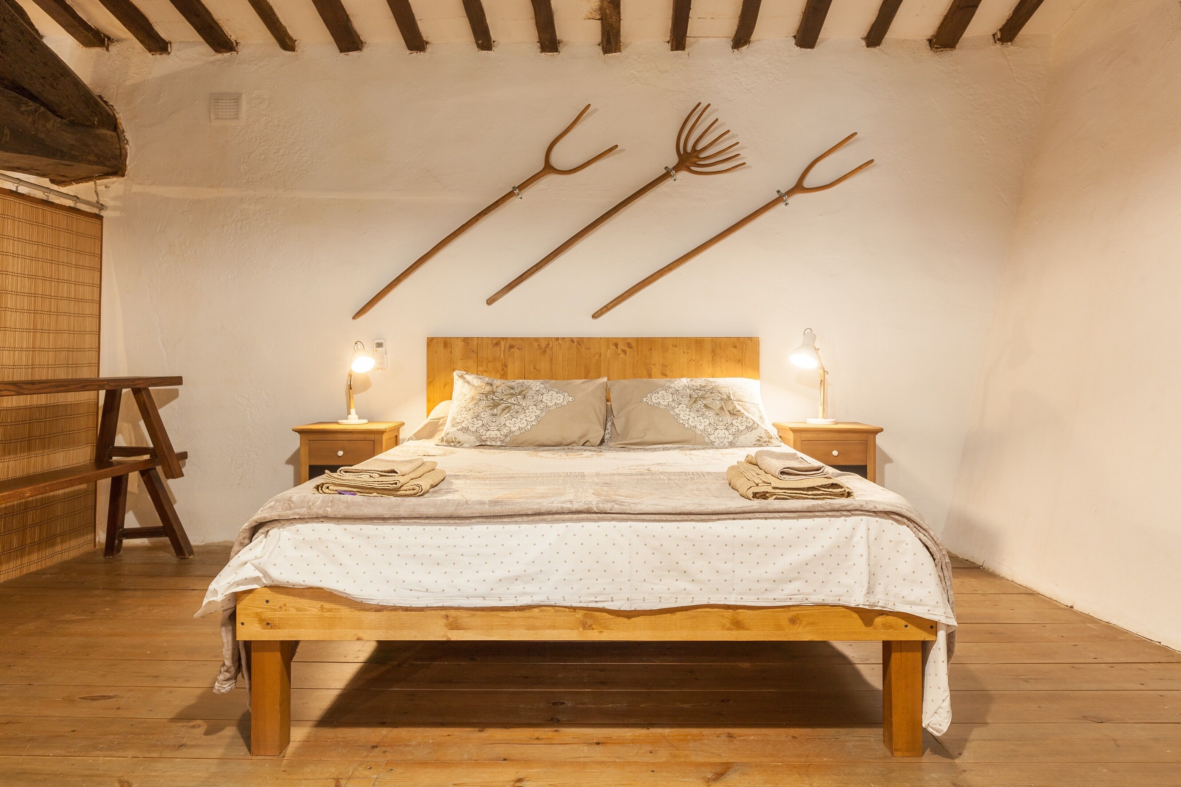 Una cama de madera con una colcha blanca, dos mesitas de noche con lámparas, y una escalera de madera apoyada en la pared.