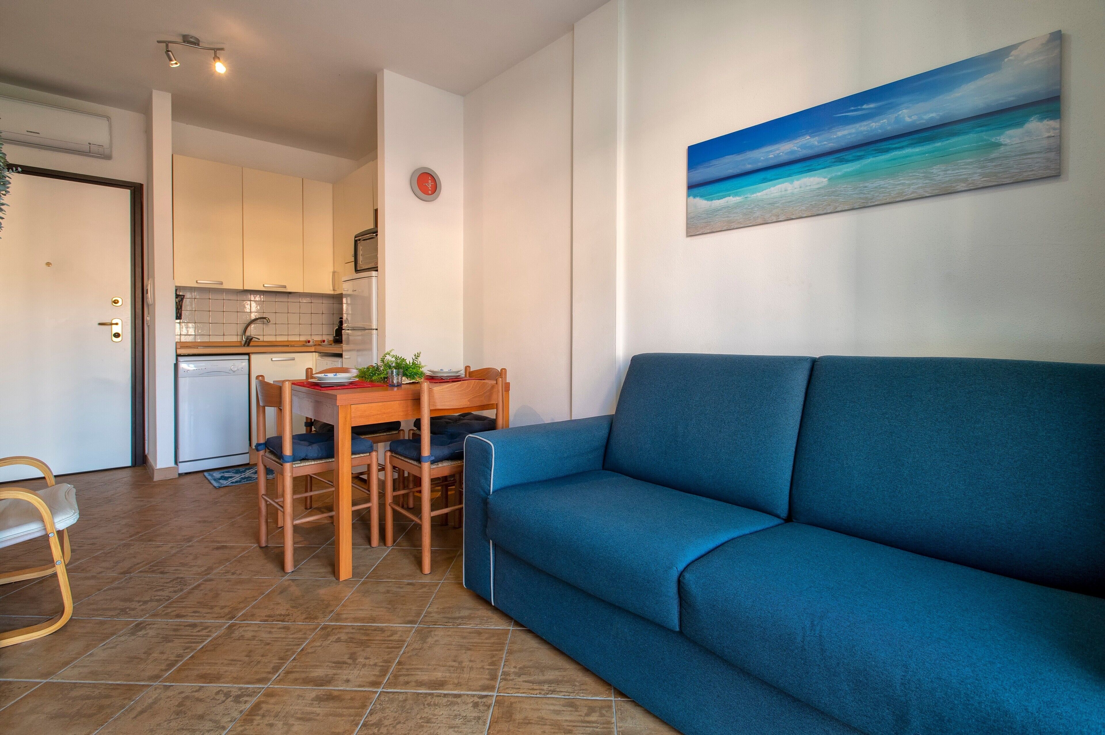 Una sala de estar con un sofá azul, una mesa de comedor con sillas, una cocina con armarios blancos y un cuadro con una playa en la pared.