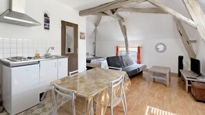 Una cocina compacta con estufa, pileta y muebles bajos blancos. Hay una mesa de comedor con sillas y una pequeña mesa de madera con un televisor. La habitación tiene vigas de madera a la vista y una cama con una colcha gris.