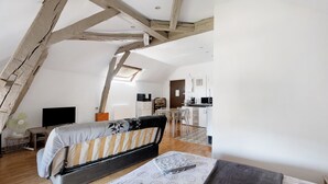 Un dormitorio estilo loft con vigas de madera a la vista, un sofá cama, un escritorio con silla, un pequeño ventilador y una kitchenette al fondo.