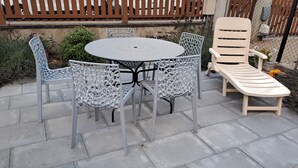 En terrasse med et rundt bord og fire stole, en liggestol og et træhegn i baggrunden.