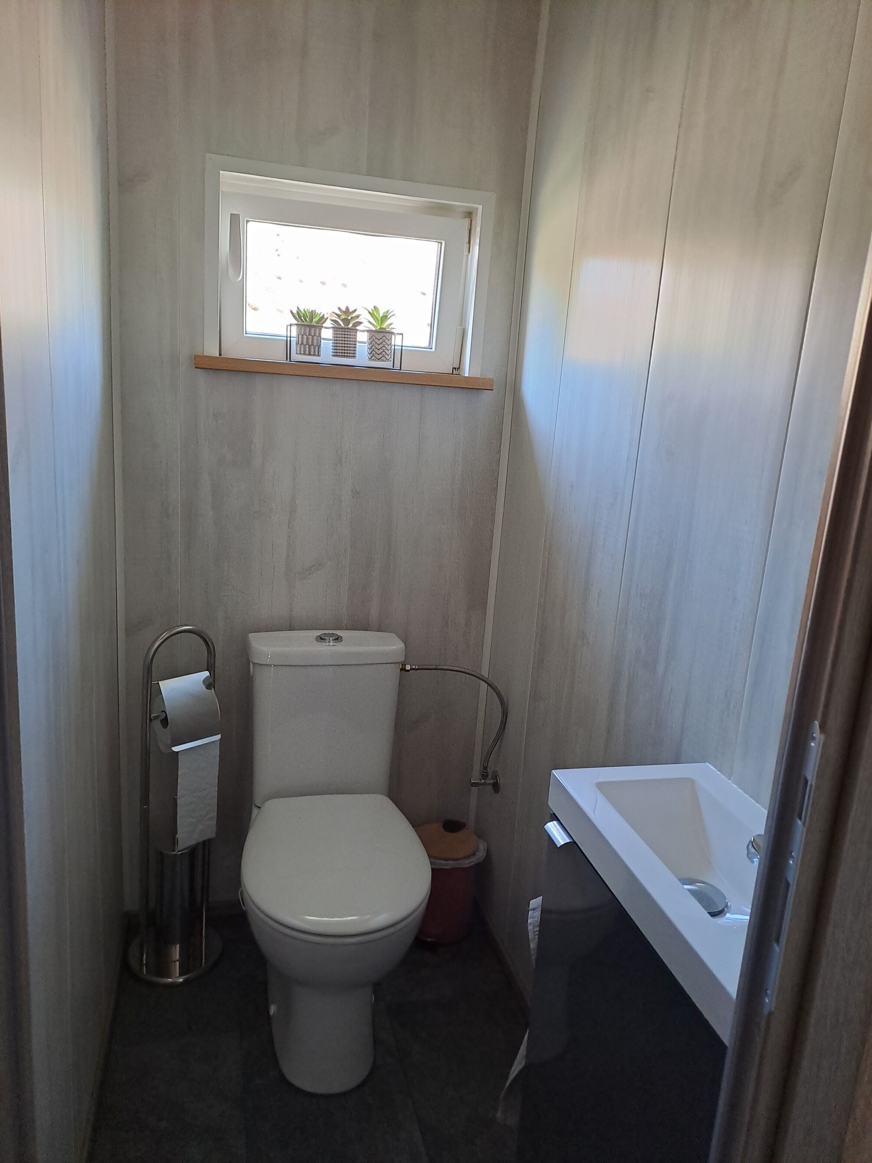 A compact bathroom with a toilet, a small window with plants, a shelf above the toilet, and a bathtub.