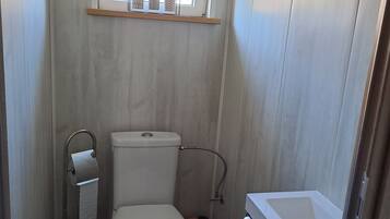 A compact bathroom with a toilet, a small window with plants, a shelf above the toilet, and a bathtub.