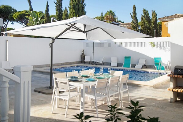 A poolside dining area with a table set for four, a covered seating area, and a pool with a blue mosaic border.
