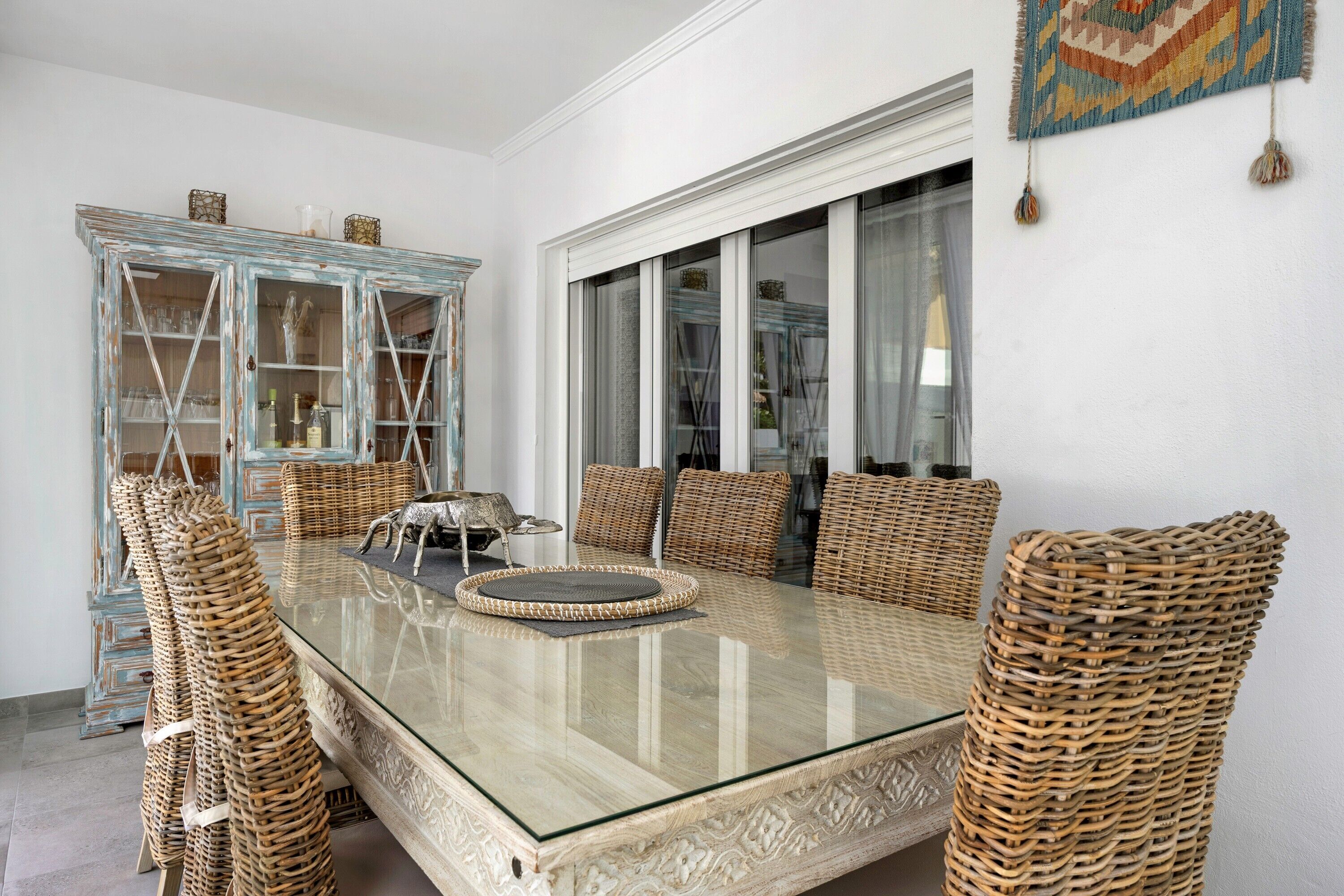 A dining area with a glass-top table, wicker chairs, and a blue-framed cabinet.