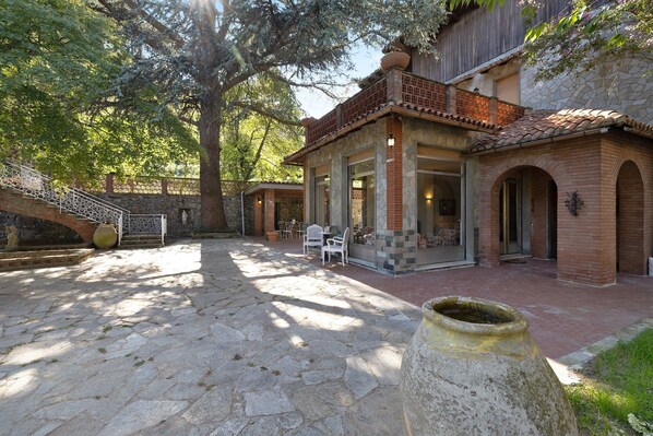 Un patio con suelo de piedra, un árbol grande, una zona de estar con sillas y un edificio de arquitectura de ladrillo y piedra.