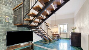 Un escalier avec des marches en bois et des rampes en métal, un téléviseur à écran plat posé sur un meuble en bois, un coin repas avec une table et des chaises, et une fenêtre laissant entrer la lumière naturelle.