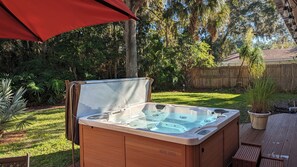A hot tub in a backyard with a wooden deck, surrounded by greenery and a wooden fence.