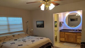 Un dormitorio con cama, ventilador de techo, mesita de noche, lámpara, y un baño con lavabo, espejo y retrete.